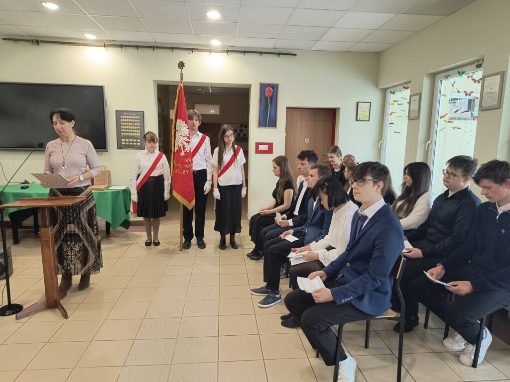 Podczas uroczystości ślubowania absolwentów, kobieta przy mównicy wygłasza przemówienie skierowane do zgromadzonych uczniów. Obecność pocztu sztandarowego oraz skupiona postawa uczestników podkreślają doniosły i formalny charakter tego ważnego etapu zakończenia edukacji.