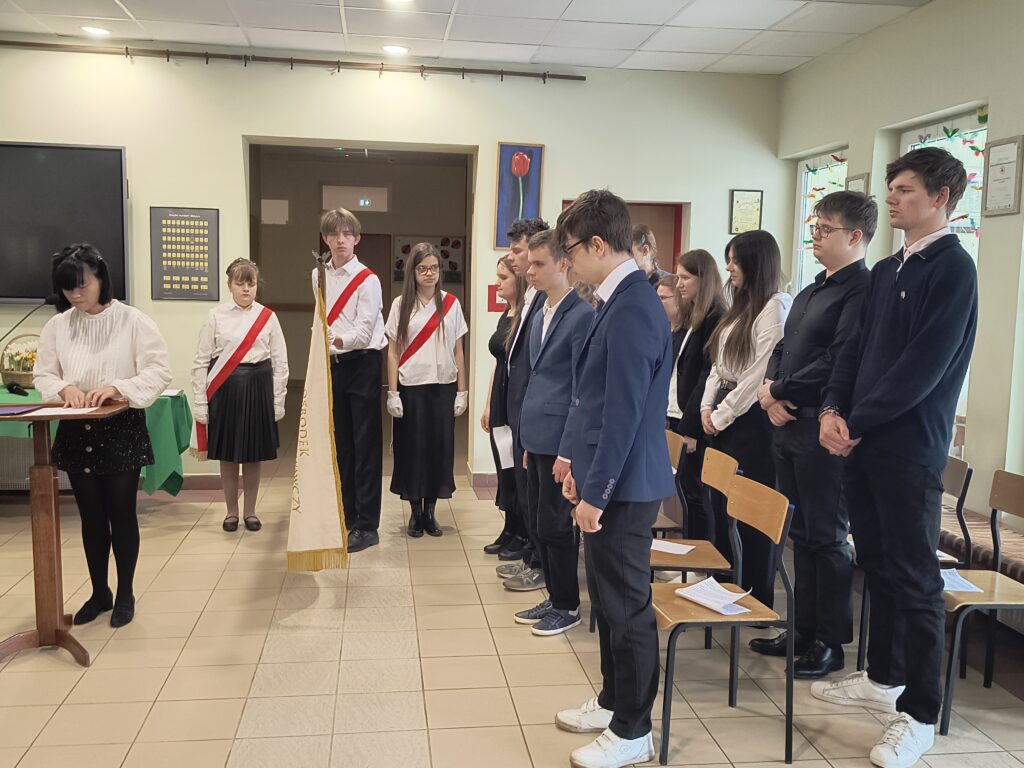 Na zdjęciu uwieczniono uroczystą akademię, podczas której uczennica przy mównicy prowadzi ceremonię w obecności pocztu sztandarowego. Zgromadzeni obok uczniowie w strojach galowych stoją w skupieniu, uczestnicząc w tym podniosłym momencie zakończenia edukacji.
