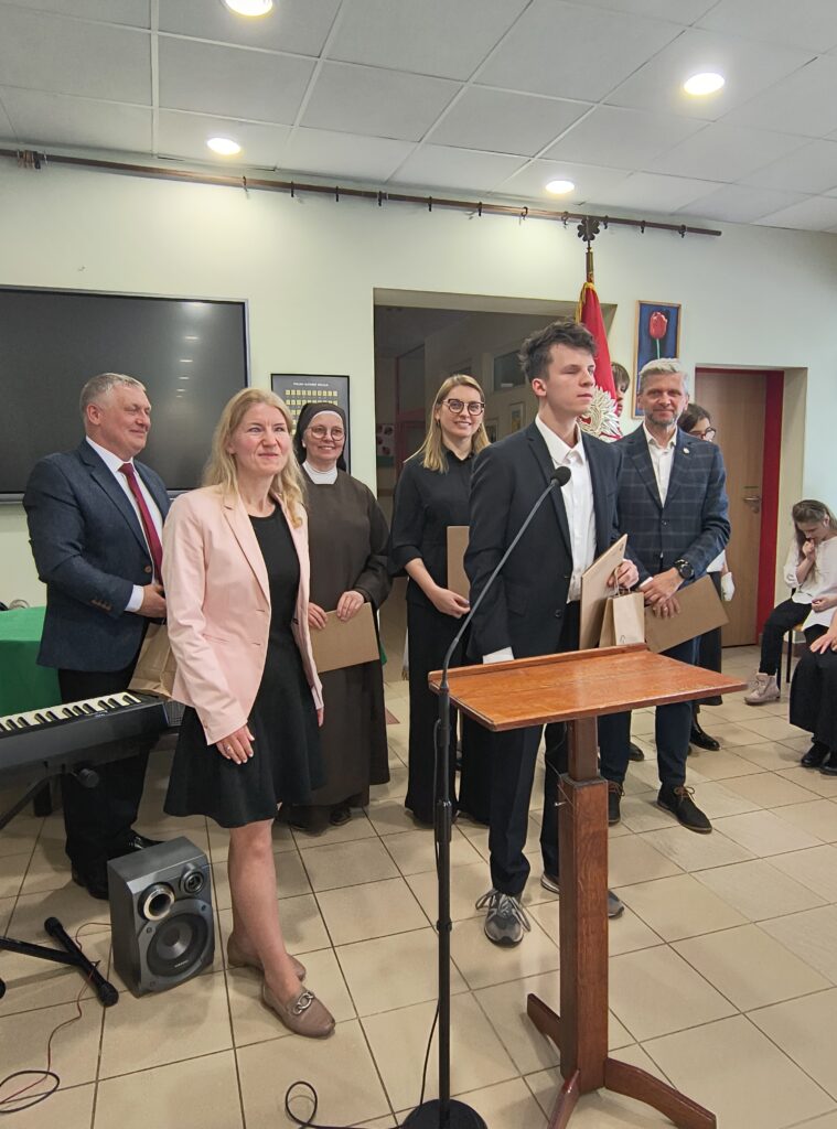 Na fotografii uwieczniono uroczysty moment wyróżnienia jednego z absolwentów podczas akademii w Ośrodku w Laskach. Młody mężczyzna w garniturze stoi przy mównicy, trzymając w rękach dokumenty oraz upominek, a za jego plecami towarzyszą mu dumni nauczyciele oraz pracownicy placówki. Cała scena podkreśla podniosły charakter wydarzenia oraz uznanie dla osiągnięć uczniów na zakończenie etapu ich edukacji.