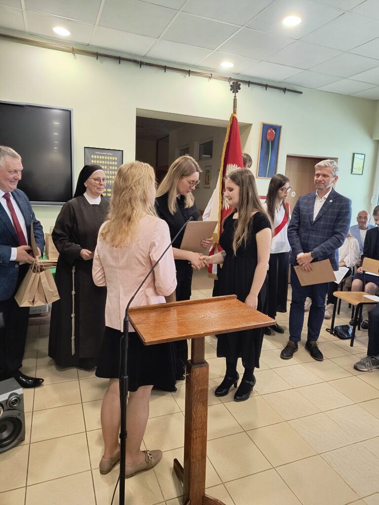 Te fotografie stanowią dopełniające się ujęcia uroczystości: na pierwszej z nich widzimy oficjalne przemówienie rozpoczynające ceremonię, podczas gdy druga uwiecznia wzruszający moment, w którym absolwentka odbiera świadectwo i gratulacje z rąk kadry ośrodka. Obrazy te tworzą pełną relację z przebiegu tego ważnego wydarzenia, od części oficjalnej po indywidualne wyróżnienia uczniów.