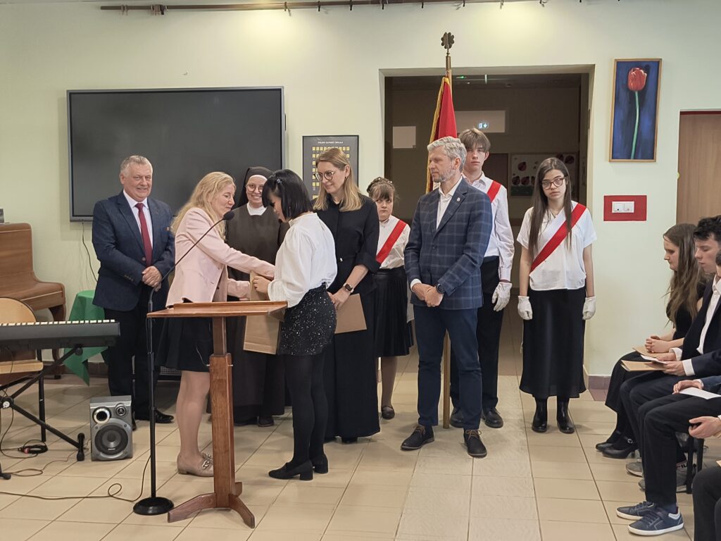 Na tym zdjęciu uwieczniono uroczysty moment wręczania wyróżnienia lub upominku podczas szkolnej akademii. Kobieta przy mównicy przekazuje przedmiot uczennicy, która stoi przed nią, podczas gdy pozostali uczestnicy – nauczyciele oraz obserwujący ceremonię uczniowie – z uwagą przyglądają się tej chwili, co podkreśla podniosły i oficjalny charakter zakończenia etapu edukacji