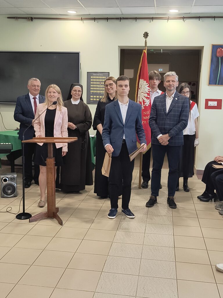 Podczas uroczystej akademii szkolnej uczeń odbiera gratulacje przy mównicy w obecności kadry nauczycielskiej oraz pocztu sztandarowego, co podkreśla doniosły charakter wydarzenia.
