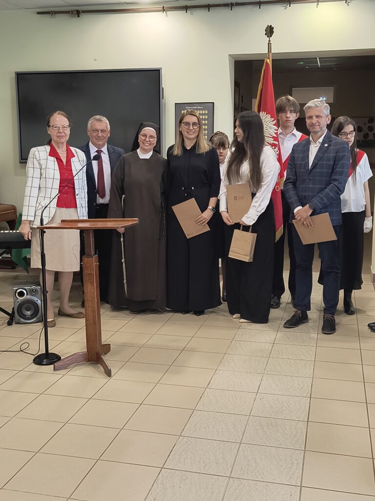 Podczas uroczystej akademii szkolnej widać grupę nauczycieli i dumnych uczniów.