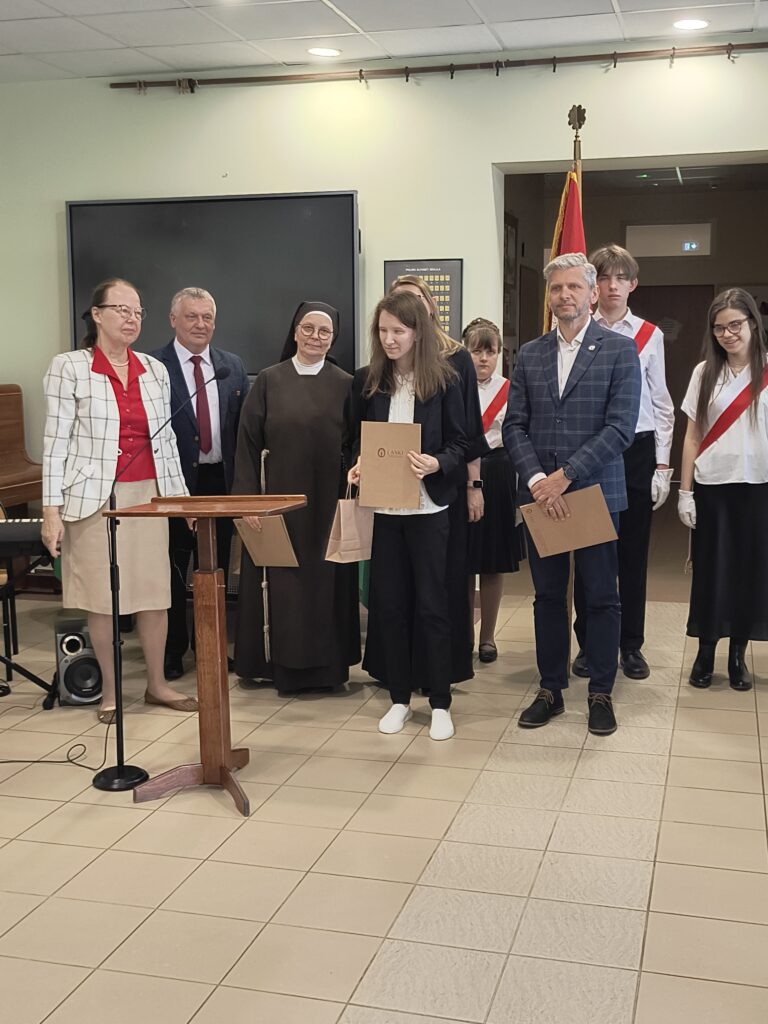 Podczas uroczystej akademii szkolnej widać grupę nauczycieli i dumnych uczniów. Kolejna uczennica odebrała nagrodę.