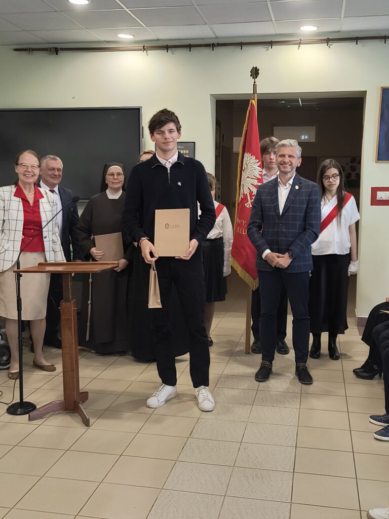 Podczas uroczystej akademii szkolnej widać grupę nauczycieli i dumnych uczniów. Uczeń technikum odebrała nagrodę.