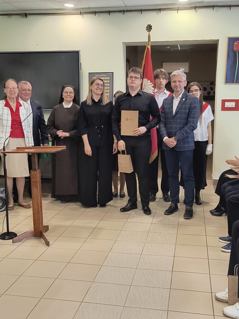 Podczas uroczystej akademii szkolnej widać grupę nauczycieli i dumnych uczniów. Drugi uczeń technikum odebrała nagrodę.