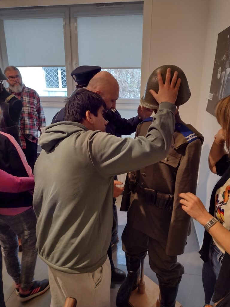 Uczniowie z zaciekawieniem oglądają i dotykają historyczny mundur oraz hełm milicyjny prezentowany na manekinie.
