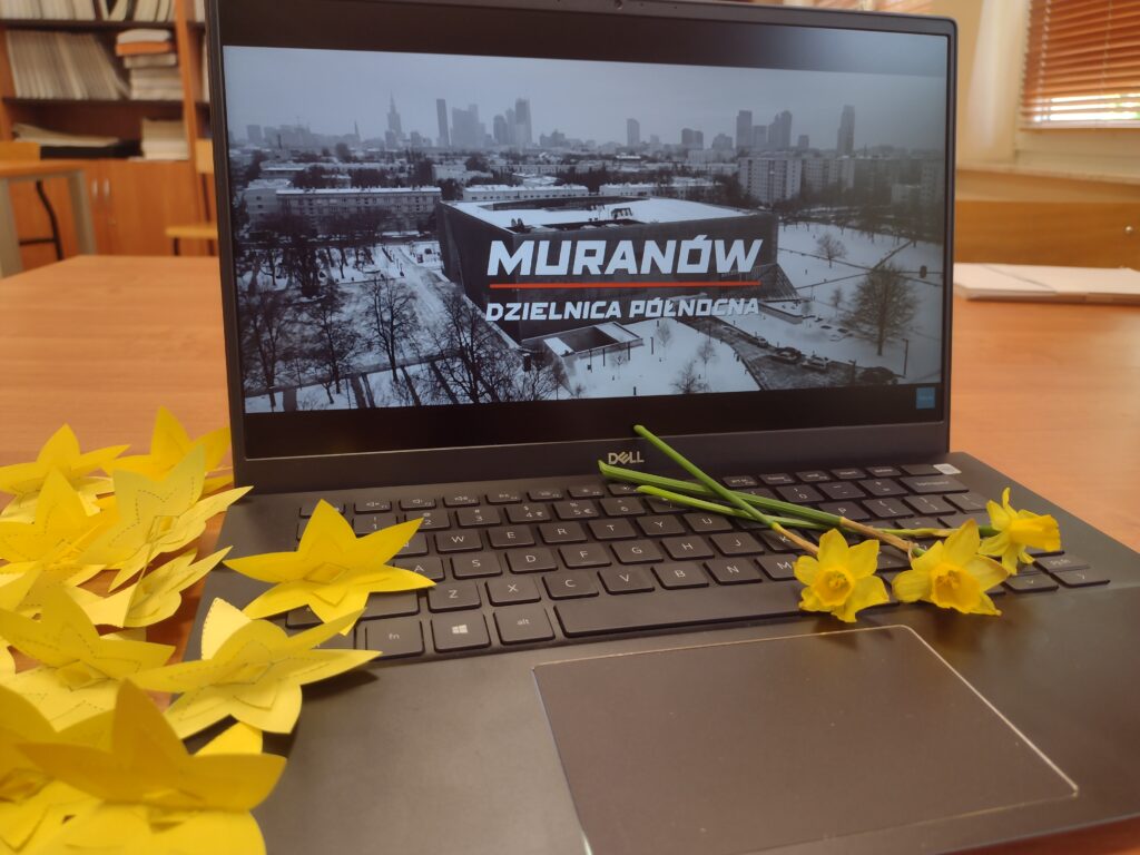 Zdjęcie przedstawia symboliczną kompozycję papierowych i żywych żonkili ułożonych przy laptopie z wyświetlonym filmem „Muranów – Dzielnica Północna”, co obrazuje udział w akcji społeczno-edukacyjnej upamiętniającej 83. rocznicę powstania w getcie warszawskim.