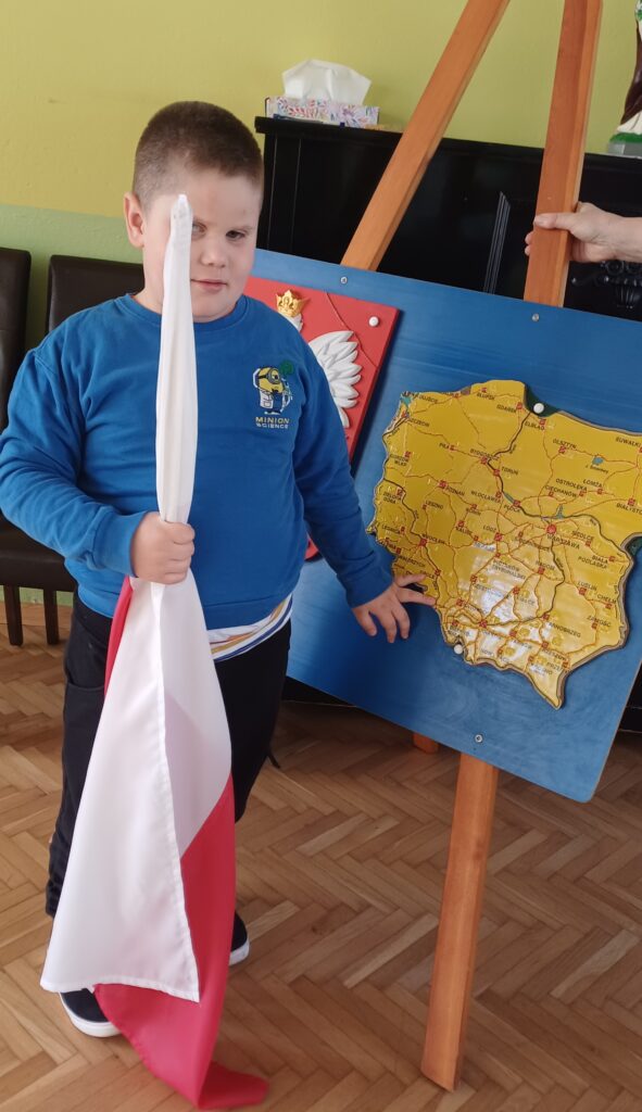 Chłopiec trzyma flagę Polski. W tle Godło i mapa Polski.