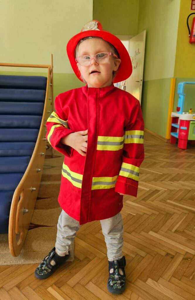 Stojący chłopiec w kurtce i hełmie strażackim.