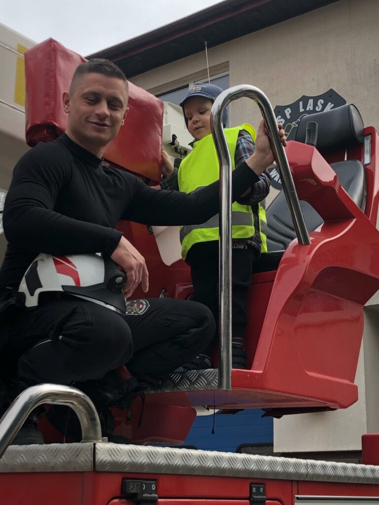 Chłopiec w kamizelce odblaskowej stoi przed  siedziskiem umieszczonym na  wozie strażackim, obok strażak