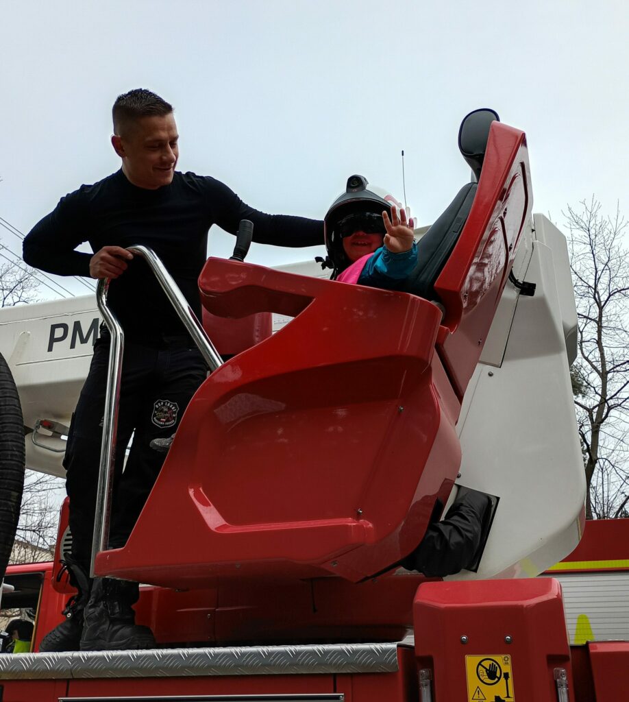 Dziewczynka w kasku i osłonie na oczy, siedzi w fotelu umieszczonym na wozie strażackim, wyciąga rękę do przodu, obok niej stoi strażak 