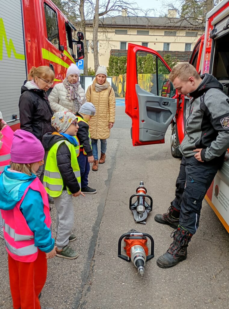 Dzieci i opiekunki stoją naprzeciwko pana strażaka, między nimi na ziemi leżą dwa duże narzędzia 