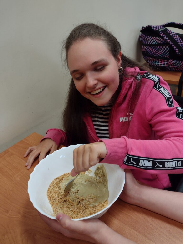 Uśmiechnięta uczennica w różowej rozpinanej bluzie z zaangażowaniem miesza łyżką chrupiące ciasto kataifi z kremem pistacjowym w białej misce, przygotowując nadzienie do czekolady dubajskiej.