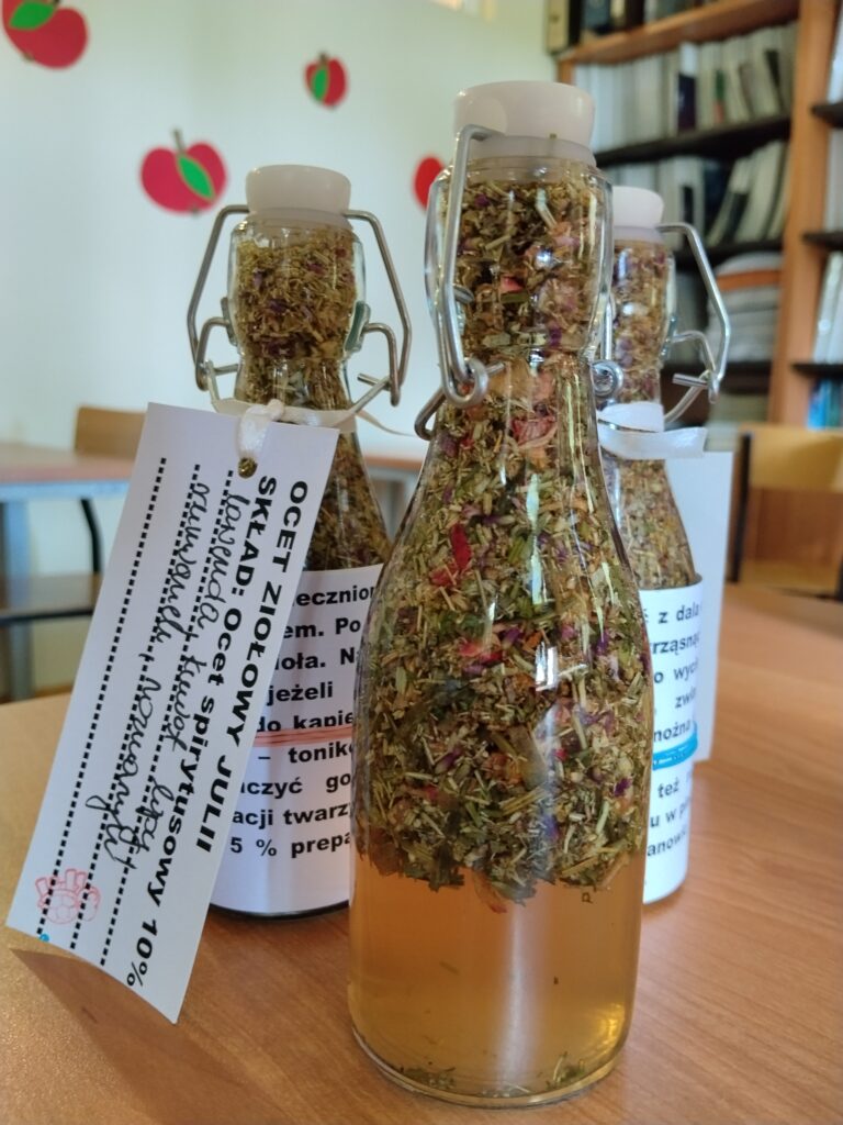 Gotowe i zamknięte w butelkach z krachlą octy ziołowe z widocznymi warstwami suszonych roślin oraz spersonalizowanymi etykietami stoją na szkolnym stole jako efekt końcowy warsztatów kosmetycznych.