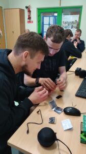 Zdjęcie przedstawia dwóch uczniów zaangażowanych w warsztaty naukowe, którzy wspólnie analizują i konfigurują niewielki mikrokontroler micro:bit. Na biurku przed nimi widoczne są elementy zestawu elektronicznego oraz peryferia komputerowe wykorzystywane do zbierania danych i trenowania modeli uczenia maszynowego.