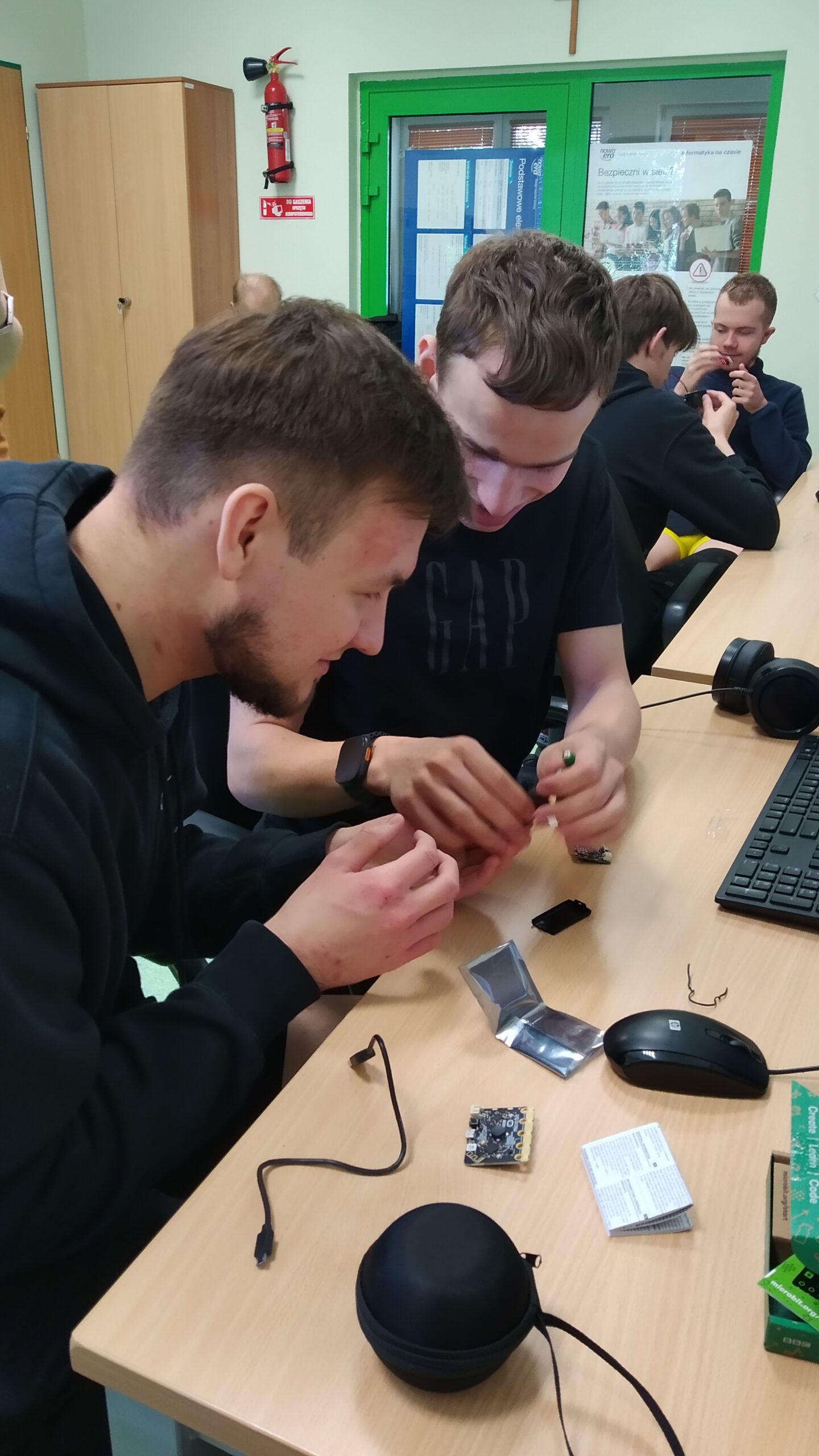 Zdjęcie przedstawia dwóch uczniów zaangażowanych w warsztaty naukowe, którzy wspólnie analizują i konfigurują niewielki mikrokontroler micro:bit. Na biurku przed nimi widoczne są elementy zestawu elektronicznego oraz peryferia komputerowe wykorzystywane do zbierania danych i trenowania modeli uczenia maszynowego.