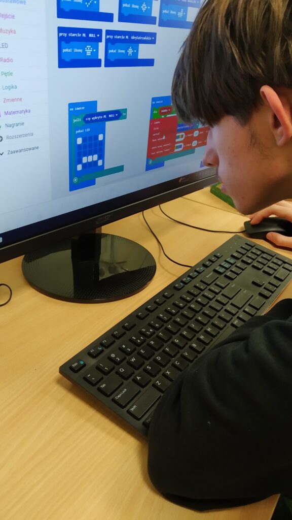 Zdjęcie przedstawia ucznia skupionego na programowaniu mikrokontrolera micro:bit za pomocą blokowego języka programowania na ekranie komputera. Tworzone bloki kodu służą do obsługi modelu uczenia maszynowego, co bezpośrednio nawiązuje do zadań z zakresu rozpoznawania gestów i sterowania dźwiękiem.