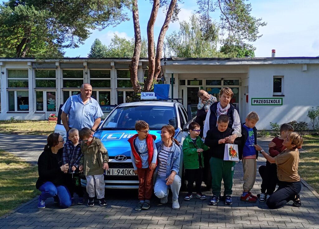 Dzieci i opiekunowie,  w pobliżu samochodu do nauki jazdy, za nimi budynek przedszkola