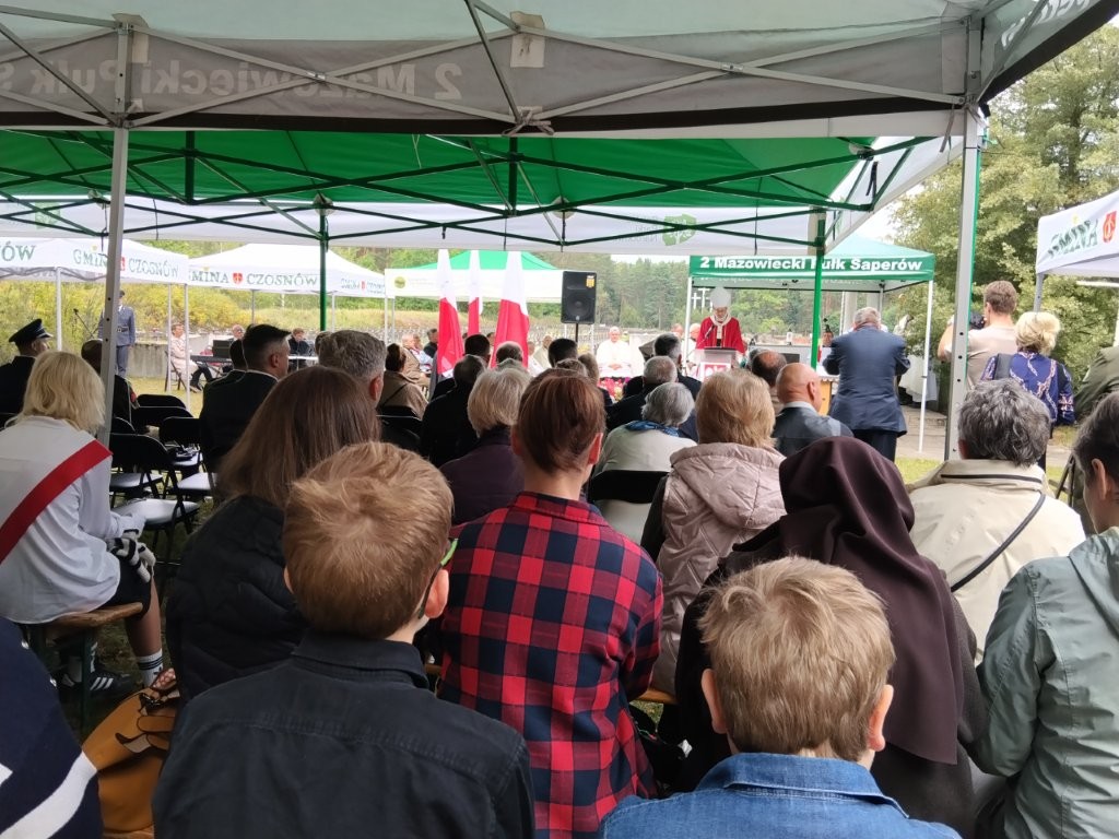 Uczestnicy uroczystości siedzą pod dużym namiotem plenerowym i słuchają przemawiającego przy ambonce duchownego, w tle widoczne są polskie flagi oraz leśne otoczenie.