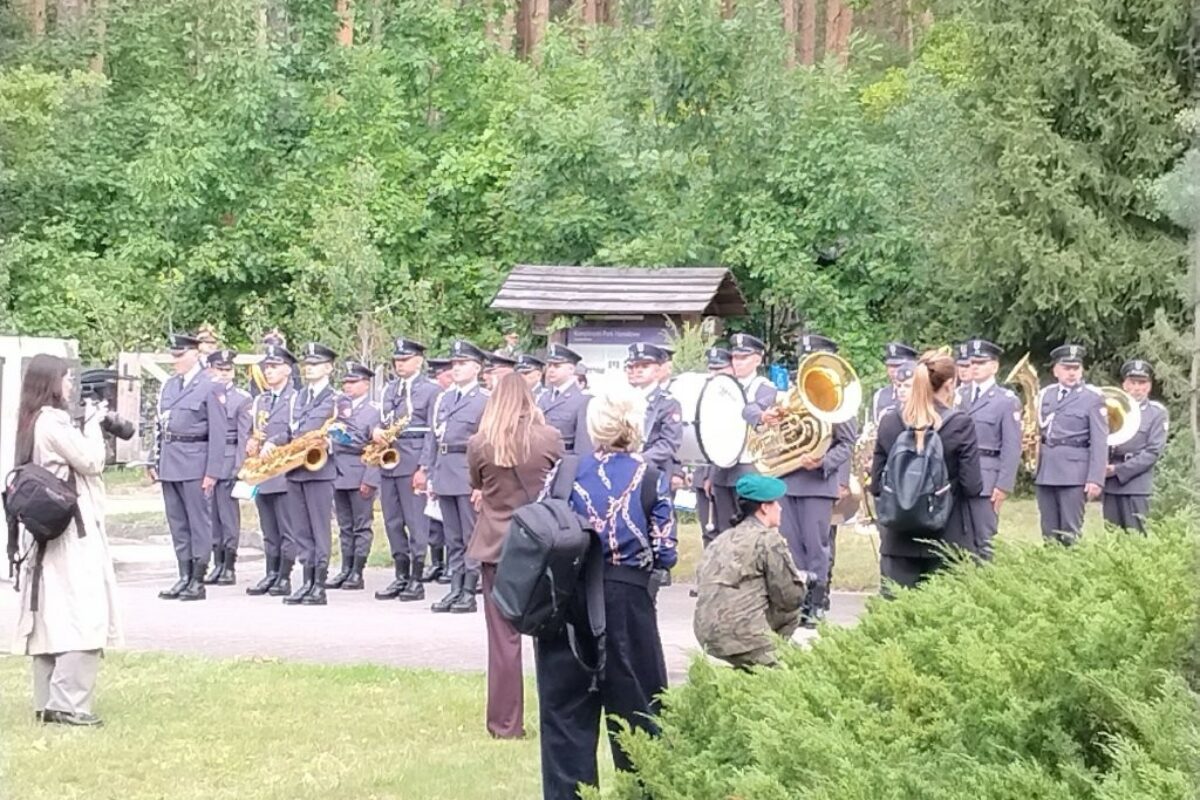 Mundurowy zespół Orkiestry Wojskowej gra na instrumentach dętych i perkusyjnych na tle lasu, podczas gdy uczestnicy uroczystości w Palmirach, stojąc w pobliżu, słuchają występów i robią zdjęcia.