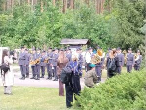 Mundurowy zespół Orkiestry Wojskowej gra na instrumentach dętych i perkusyjnych na tle lasu, podczas gdy uczestnicy uroczystości w Palmirach, stojąc w pobliżu, słuchają występów i robią zdjęcia.