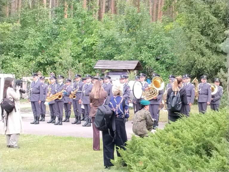 Mundurowy zespół Orkiestry Wojskowej gra na instrumentach dętych i perkusyjnych na tle lasu, podczas gdy uczestnicy uroczystości w Palmirach, stojąc w pobliżu, słuchają występów i robią zdjęcia.
