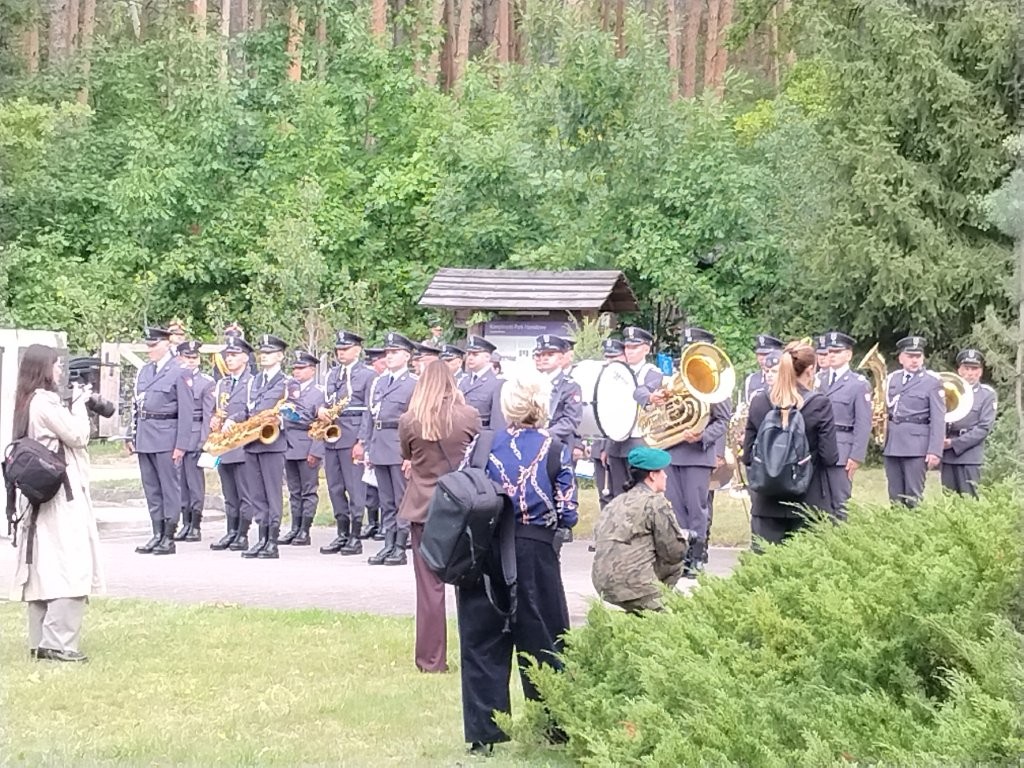 Mundurowy zespół Orkiestry Wojskowej gra na instrumentach dętych i perkusyjnych na tle lasu, podczas gdy uczestnicy uroczystości w Palmirach, stojąc w pobliżu, słuchają występów i robią zdjęcia.
