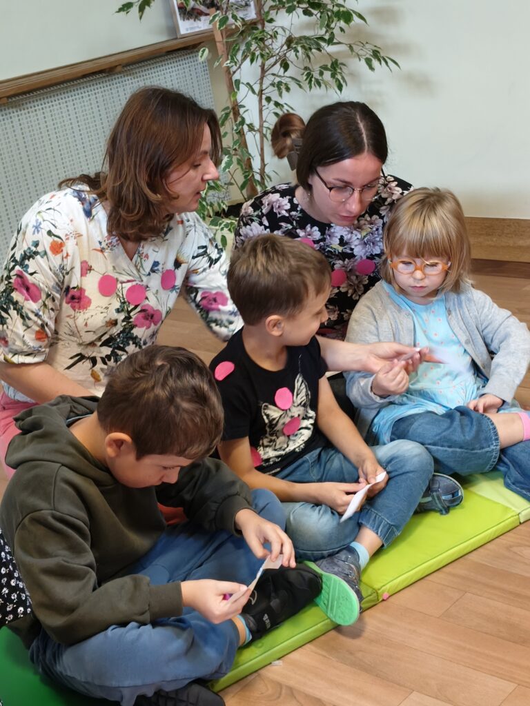 Troje uczniów z nauczycielkami siedzący na podłodze. Na bluzkach mają widoczne różowe kółeczka.