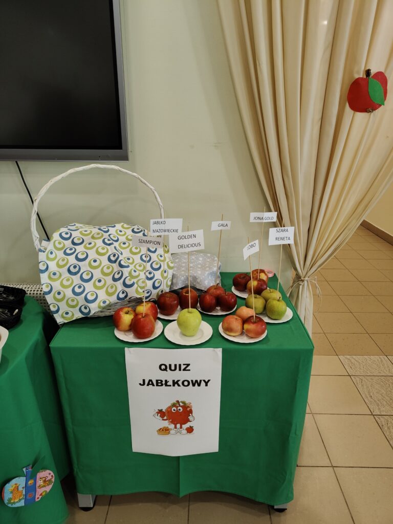Stolik przygotowany do quizu jabłkowego, na którym zaprezentowano różne odmiany jabłek podpisane osobnymi karteczkami.