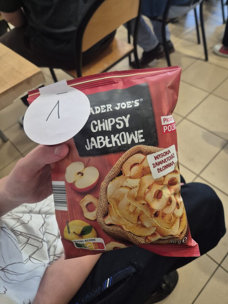 Uczestnik wydarzenia trzyma w dłoni paczkę chipsów jabłkowych oznaczoną numerkiem, wręczoną jako upominek w ramach szkolnych konkursów z okazji Światowego Dnia Jabłka.